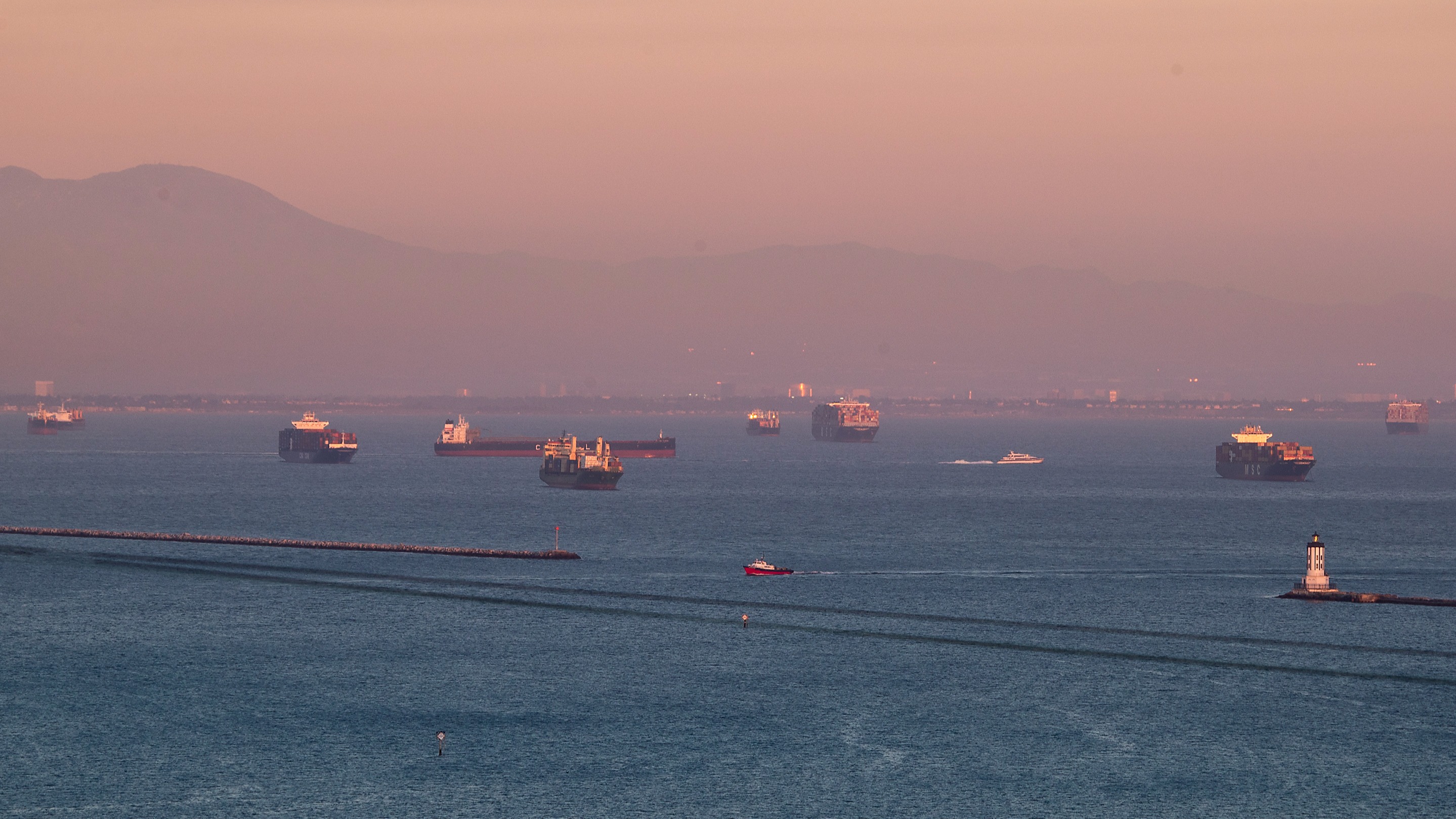 The sunset illuminates the scene of dozens of container ships siting off the coast of the Ports of Los Angeles and Long Beach, waiting to be unloaded
