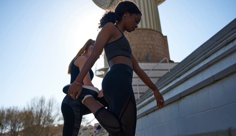Two women exercising together outdoors in the sun. The woman in the foreground is bending her leg backwards to stretch. They are both wearing all black and have their hair tied back. This photo is being used in an article answering the question