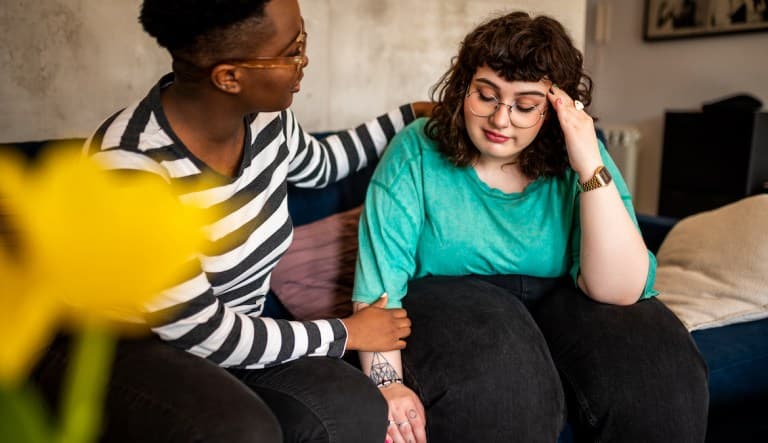Two women sitting on a cough. The woman on the right is wearing a long-sleeve striped shirt and comforting the woman on the right, who is visibly distressed and wearing a bright turquoise shirt. This photo is being used in an article about friendship inse
