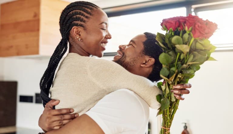 A man and woman hold each other in a loving embrace. They are both wearing white and smiling brightly at each other, and the woman is holding a bouquet of red roses. This photo is being used in an article about what happens after the honeymoon phase in a