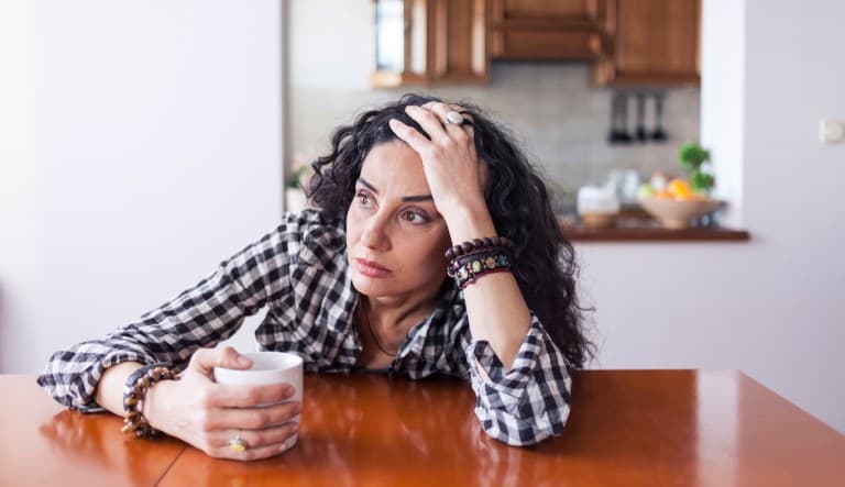 A woman sits with her head propped on one arm and her opposite hand holding onto a coffee mug. She looks forlorn as she stares off into the distance. This photo is being used to promote an article answering the question