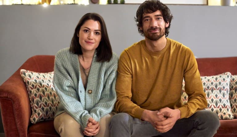 A young couple sits facing the camera with their hands clasped in front of their laps. They both have small smiles on their faces. This photo is being used in an article about what happens after the honeymoon phase in a relationship.