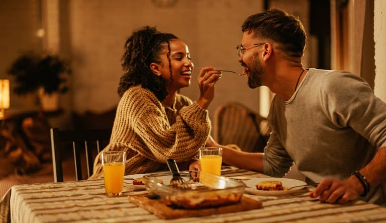 A young couple enjoy dinner at home. They are both smiling and the woman on the right is feeding the man on the left with her fork. This photo is being used in an article about what happens after the honeymoon phase in a relationship.
