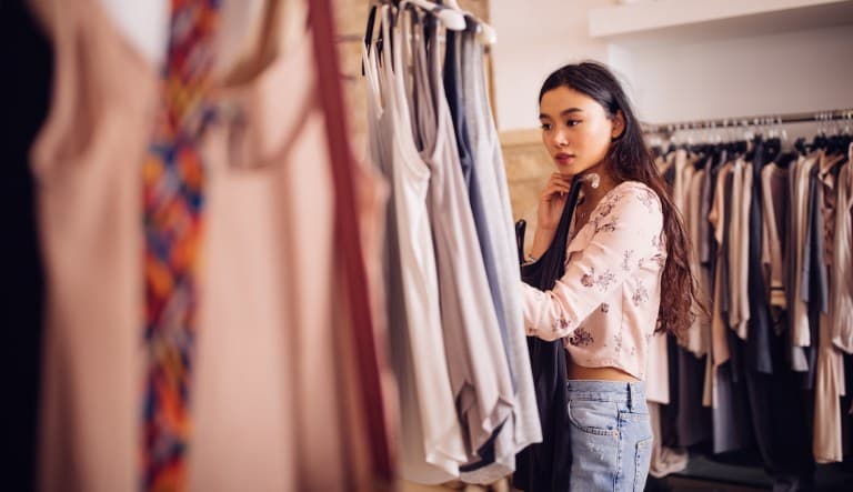 A young woman is shown browsing for clothes at a boutique. There are blurred shirts hanging in the foreground and she appears to be contemplating one shirt in particular. This photo is being used in an article about breathable fabrics.