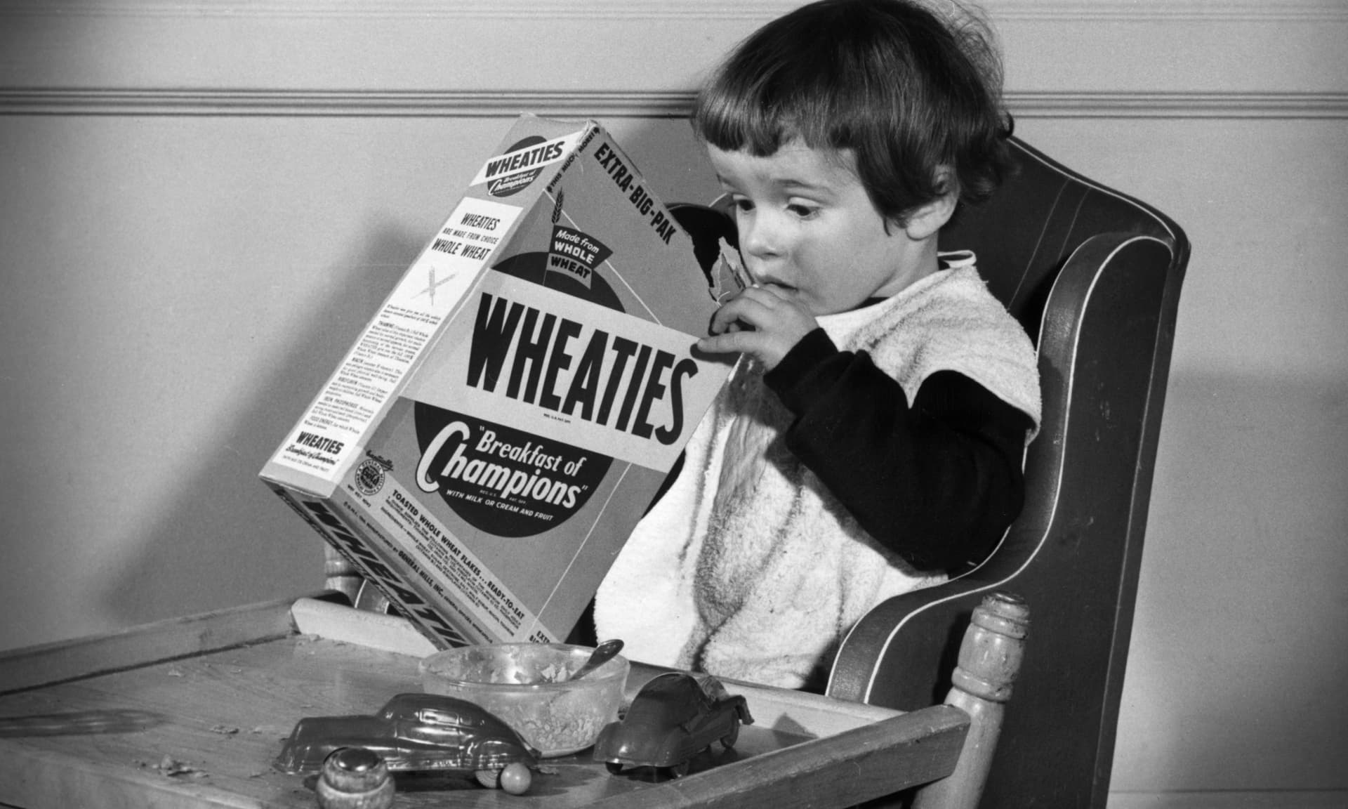Circa 1945: A young girl sits at a table, digging with one hand in a box of Wheaties breakfast cereal.
