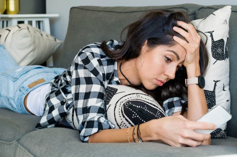 A sad woman stares at a white phone on the couch with her hand touching her head in a display of exhaustion. She is wearing a plaid shirt and light was jeans and has dark hair. This photo is being used to promote an article answering the question