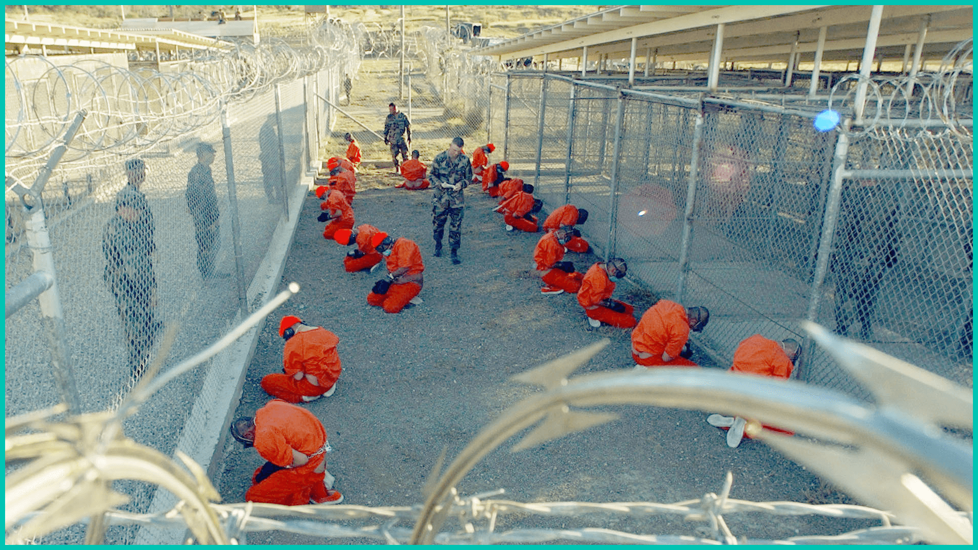 Taliban prisoners in orange jumpsuits sitingt in holding area under the watchful eyes of military police