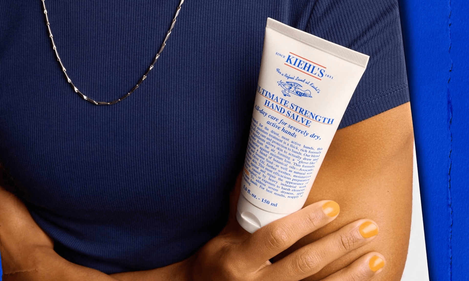 Woman in blue shirt with gold necklace holding tube of Kiehl's hand salve on blue painted background